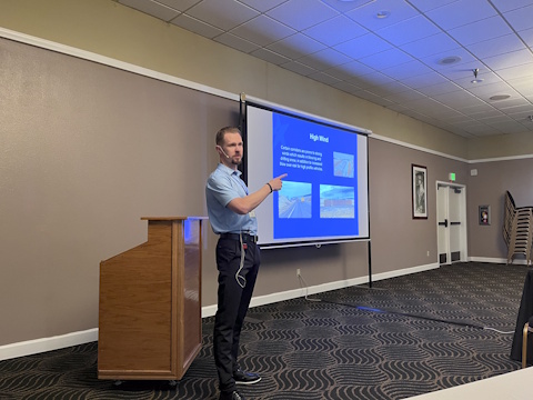 A man points with his right hand while speaking beside a projector screen.
